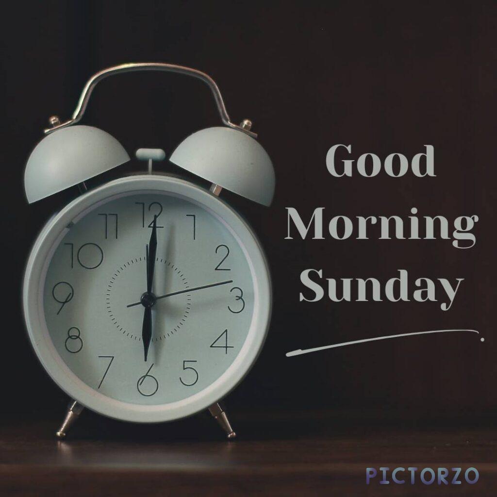 A close-up photo of a black alarm clock with the time 6 AM displayed. The words Good Morning Sunday are written in white on the clock face