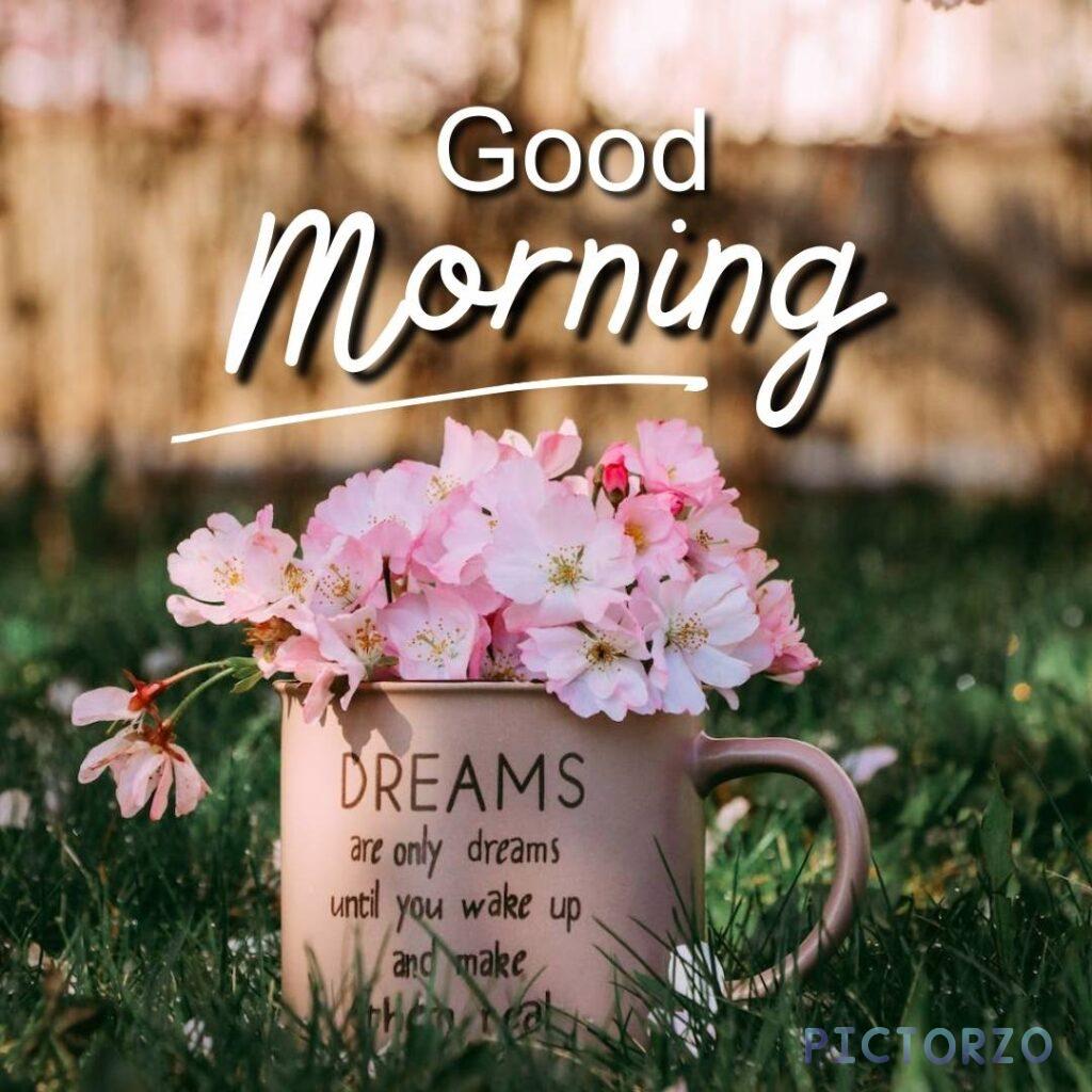 A cup filled with pink roses and other flowers sitting in the grass, with the text Good Morning and Dreams are only dreams until you wake up and make them real overlaid in the top right corner