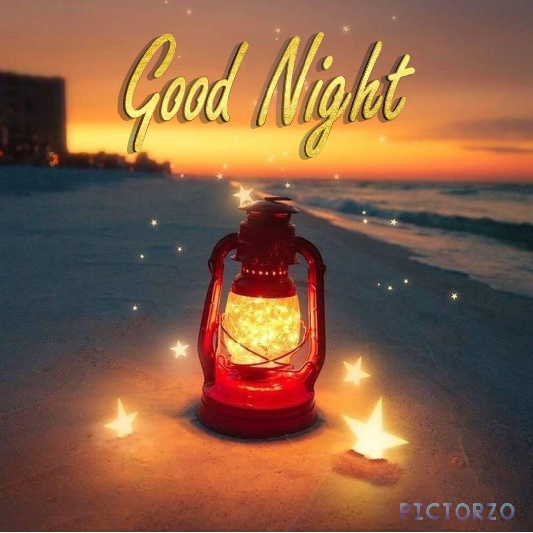 Good Night Beautiful Images 1 A close-up photo of a lantern sitting on top of a beach at night. The lantern is lit, and its warm glow casts a soft light on the surrounding sand and water. The sea is calm in the background, and the stars are visible in the sky above.