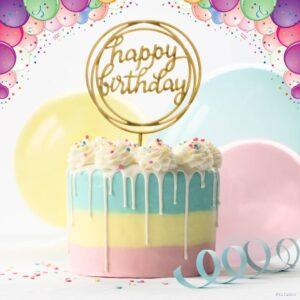 A close-up photo of a round chocolate cake with a golden happy birthday topper and balloons on a white background. The cake is decorated with white frosting, chocolate shavings, and sprinkles