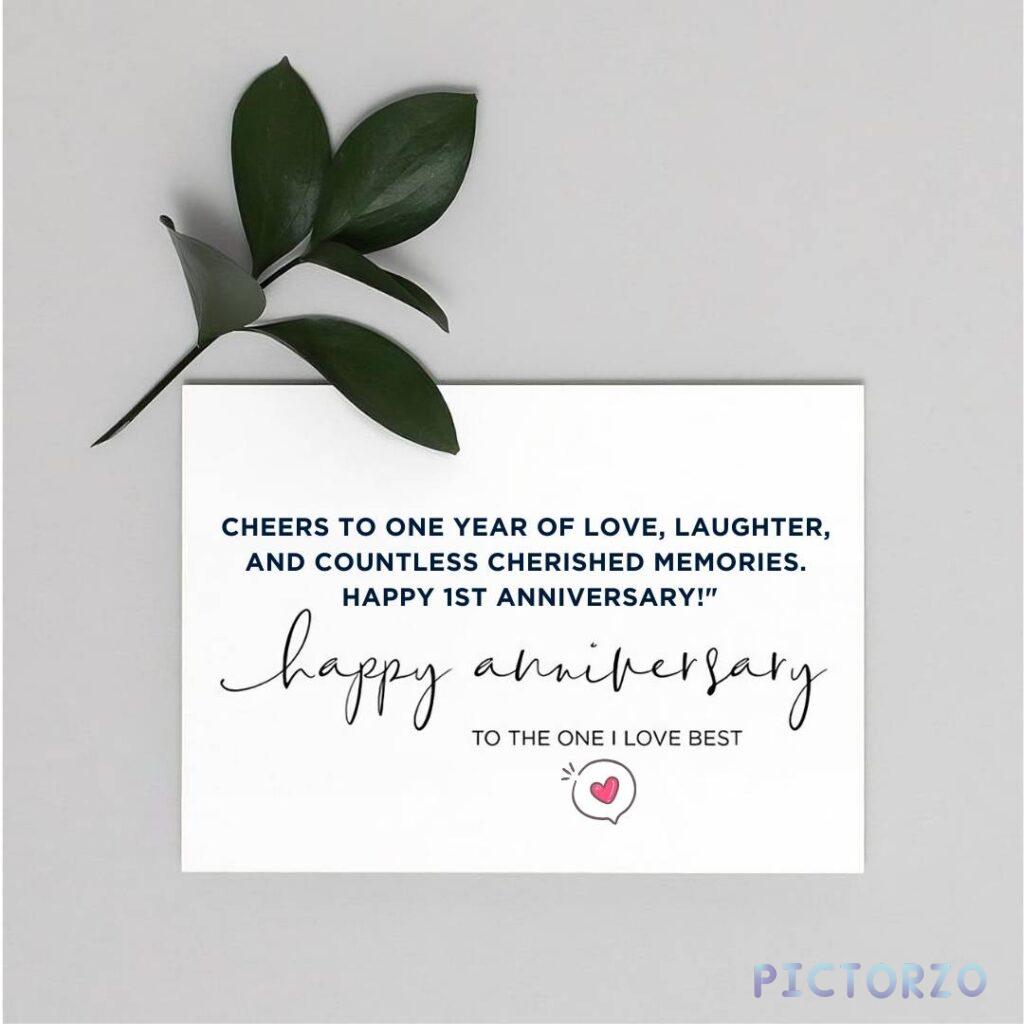 A close-up photo of a white anniversary card with a green leaf resting on top. The card has the following text written in black lettering: "Cheers to one year of love, laughter, and countless cherished memories. Happy 1st Anniversary!" The leaf has the same message written on it in smaller lettering.