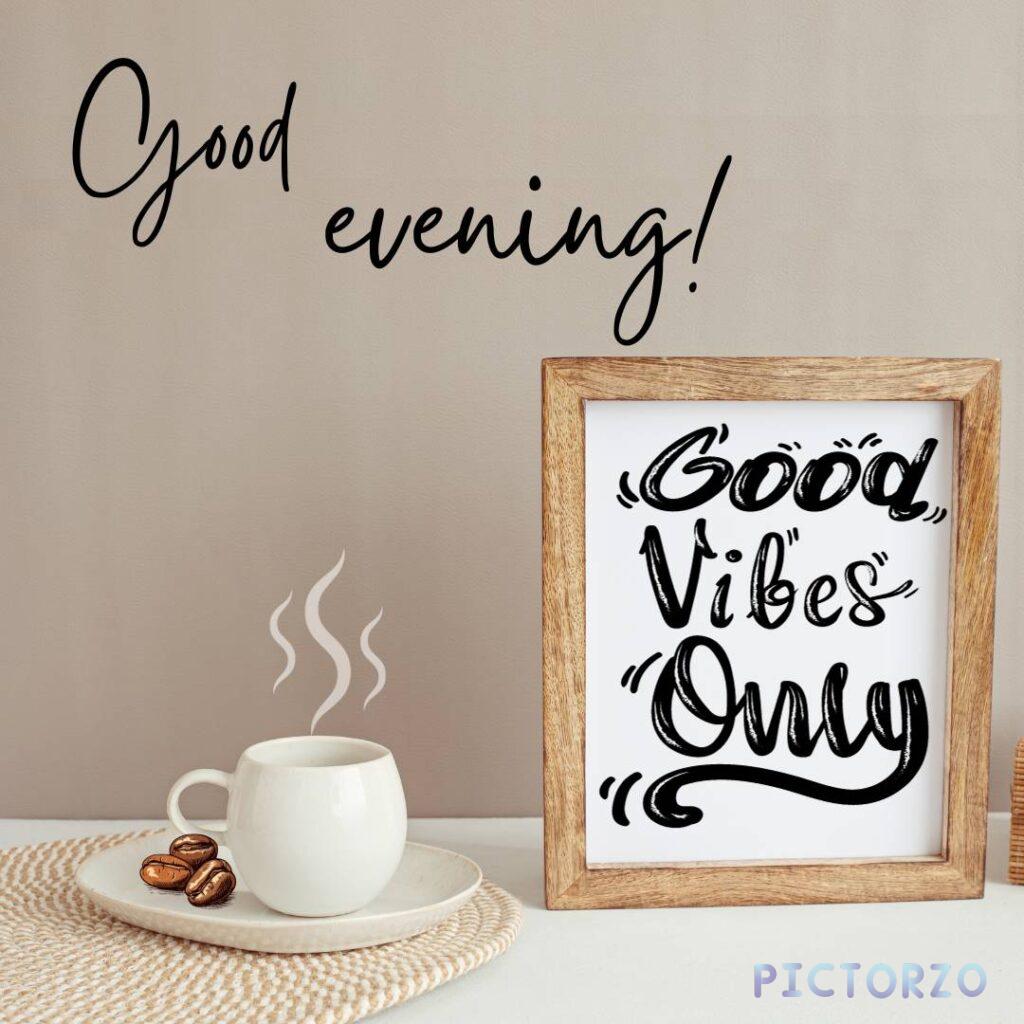 A cup of coffee sits on a wooden table next to a wooden frame with the text "Good evening! Good Vibes Only" written on it. The coffee cup is steaming and has a frothy top. The wooden frame is rustic and has a farmhouse feel to it.