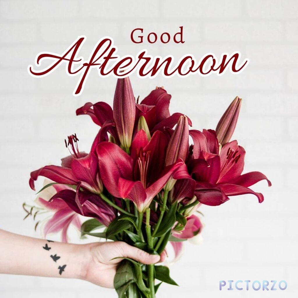 A person holding a bouquet of maroon Stargazer lilies. The lilies are in full bloom and their petals are open wide. The background is blurred, giving the focus to the flowers. The text "Good Afternoon" is superimposed on the image.