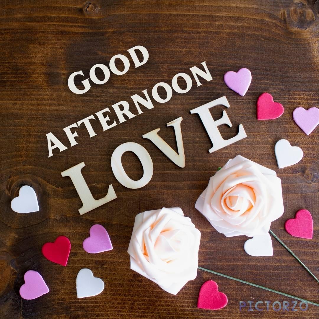 A photo of a wooden table with a bouquet of roses and a heart-shaped box of chocolates. The text "Good afternoon, love" is written on the table in front of the flowers. The image can be used to express love and affection for someone, or as a greeting card for a loved one.