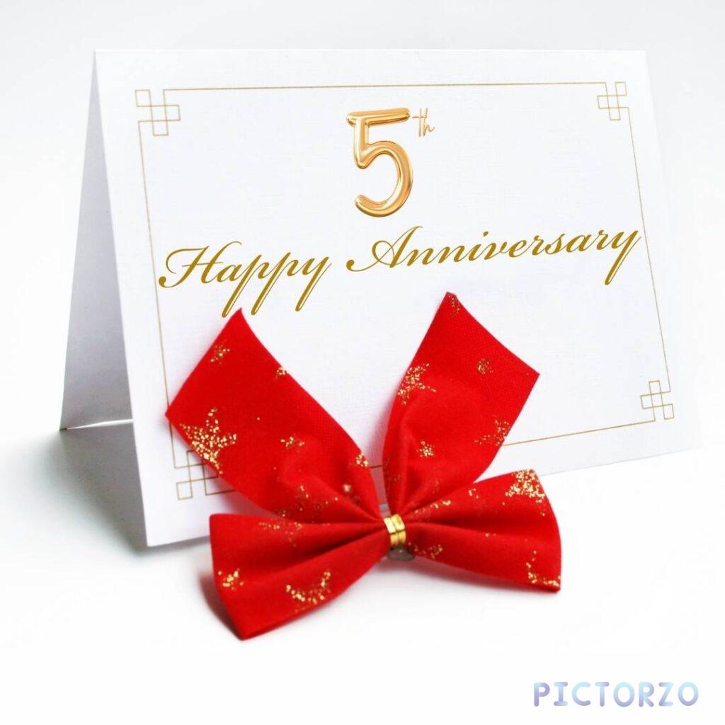 A white greeting card with a red ribbon tied in a bow. The card has the number 5 in a silver foil design and the text Happy Anniversary written in red script.