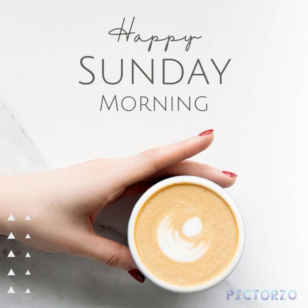 A hand with red nail polish holding a cup of coffee with latte art. The text Happy Sunday Morning is written in the top left corner.