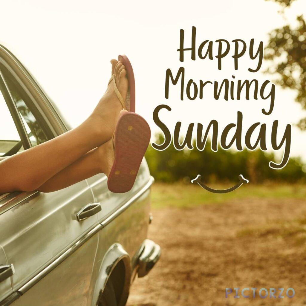 A person's legs in flip-flops are resting on the dashboard of a parked car with the windows down. The person is wearing shorts. The text "Happy Morning Sunday" is superimposed on the image with a smiley face below it.