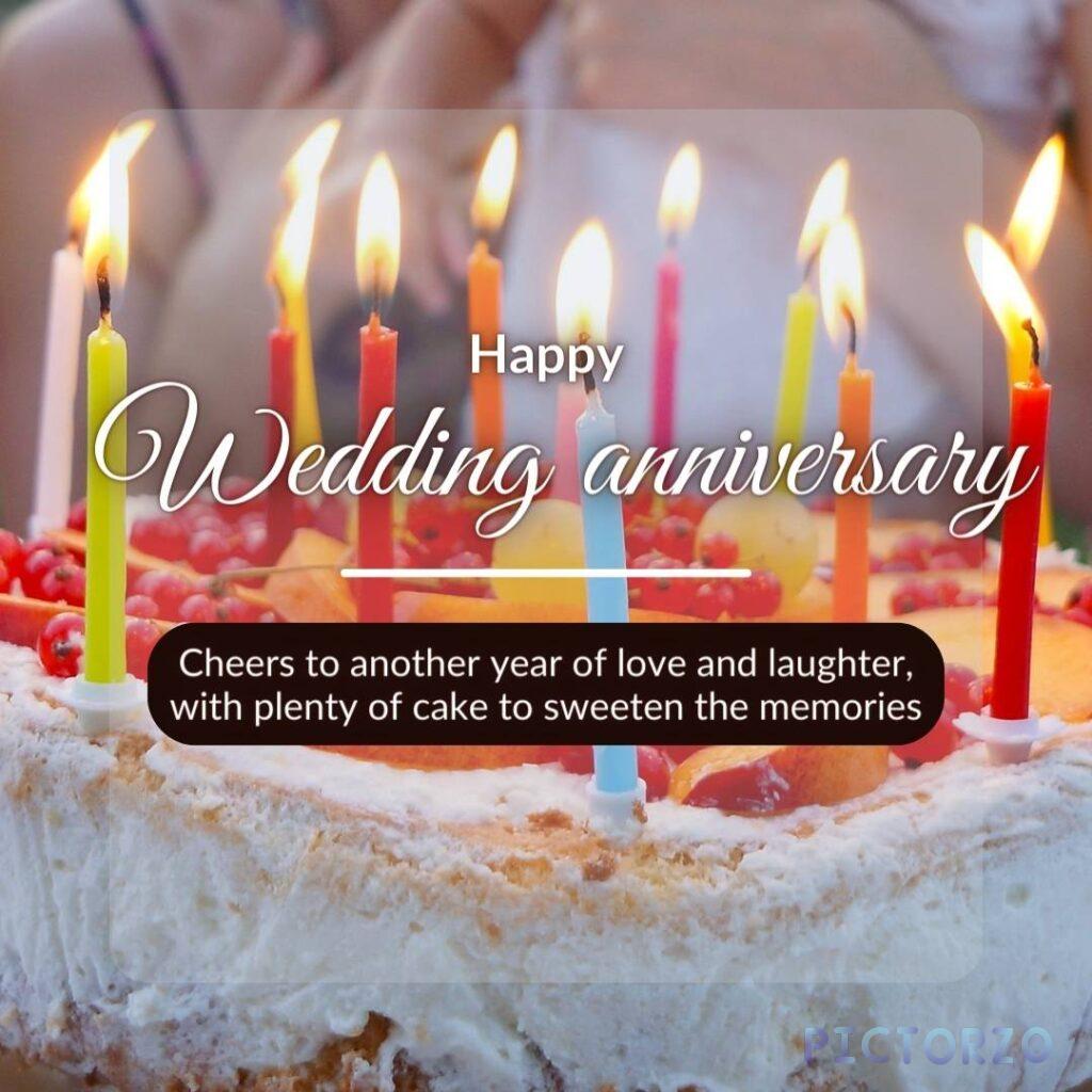 A decorated wedding cake with white frosting. The cake has lit candles and icing that reads "Happy Wedding Anniversary. Cheers to another year of love and laughter, with plenty of cake to sweeten the memories."